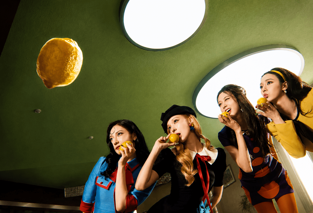 Four performers hold lemons in a retro green-yellow room from aespa’s LEMONADE music video, with a floating lemon and oval lights creating a surreal citrus-themed scene.